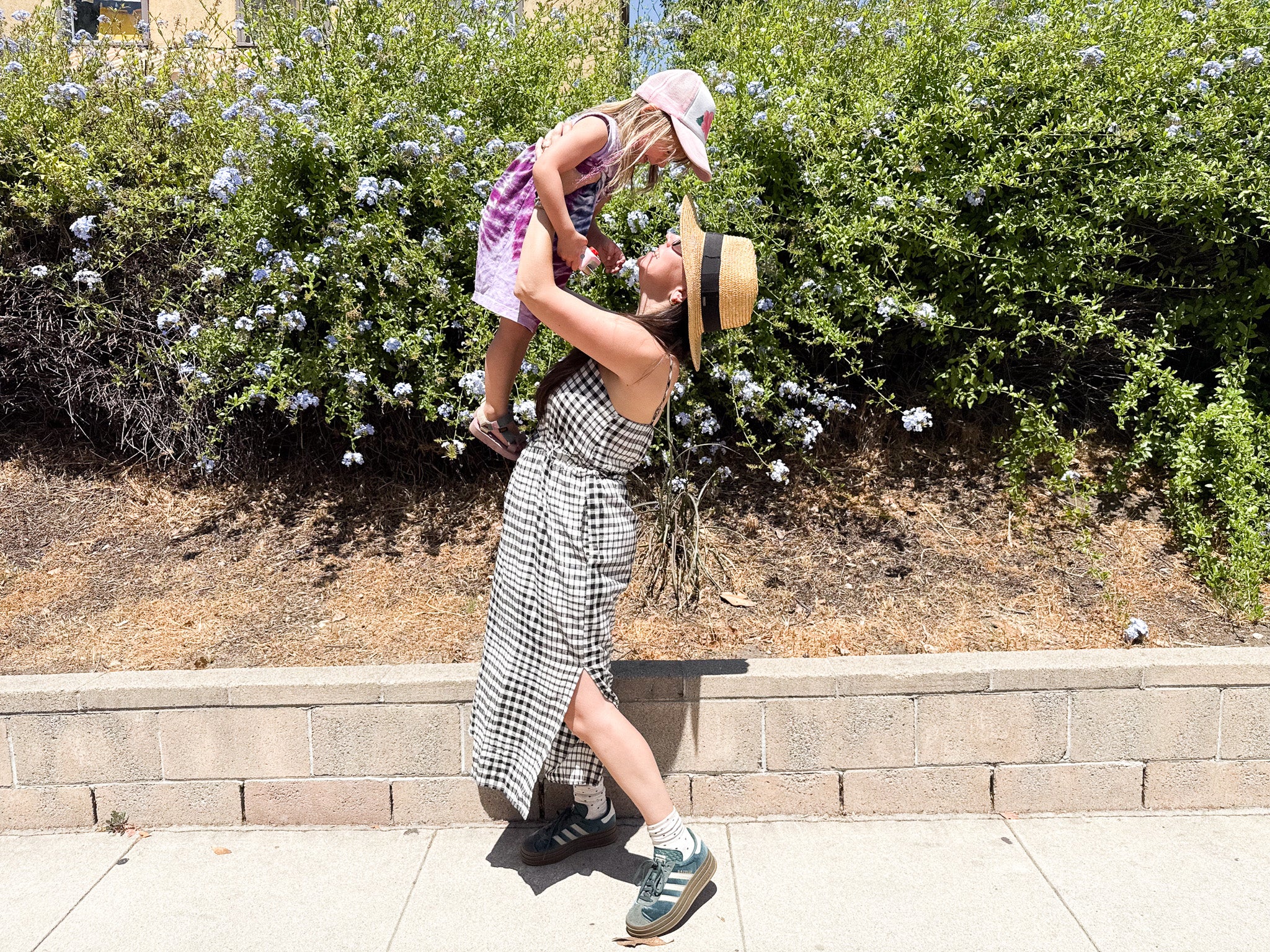 Woman in a checkered dress lifting a child in a park with greenery and flowers.