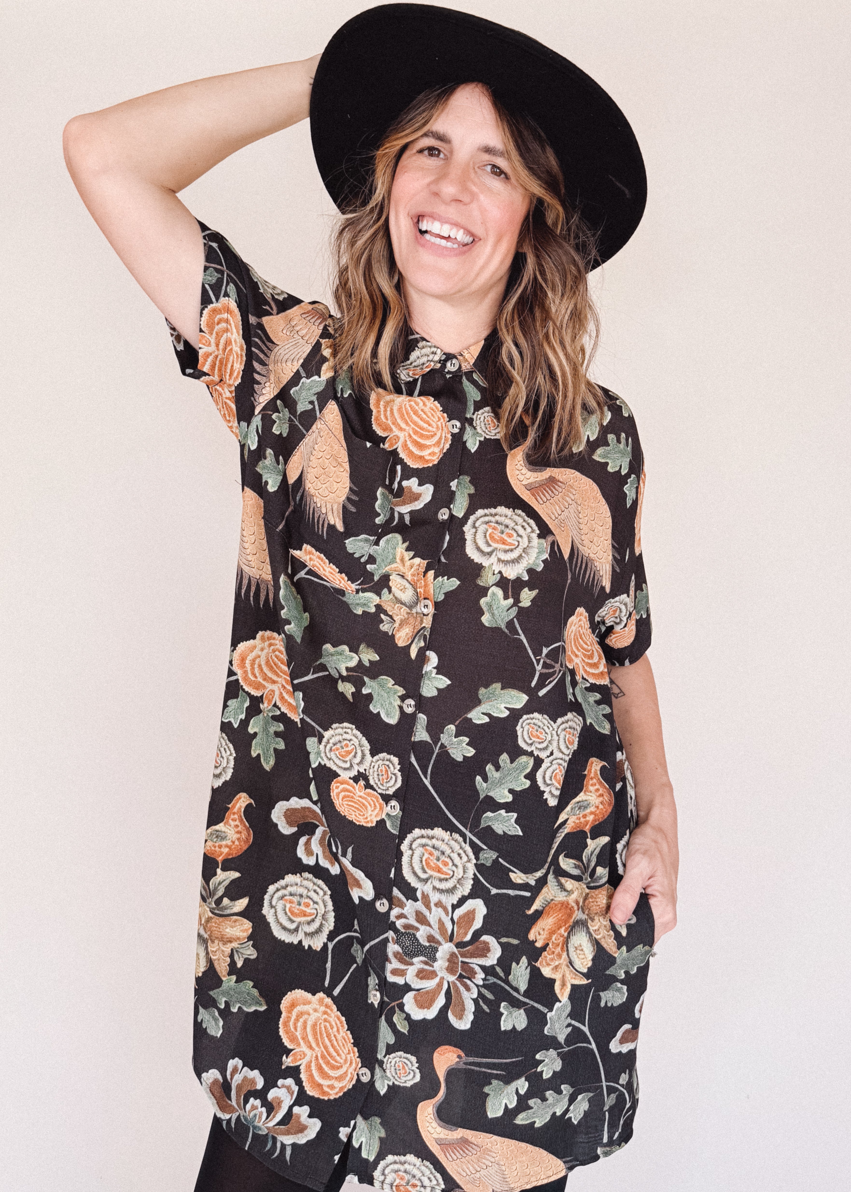 Woman wearing crane-print short sleeve button-up mini shirt dress with collar and side pockets, holding hat