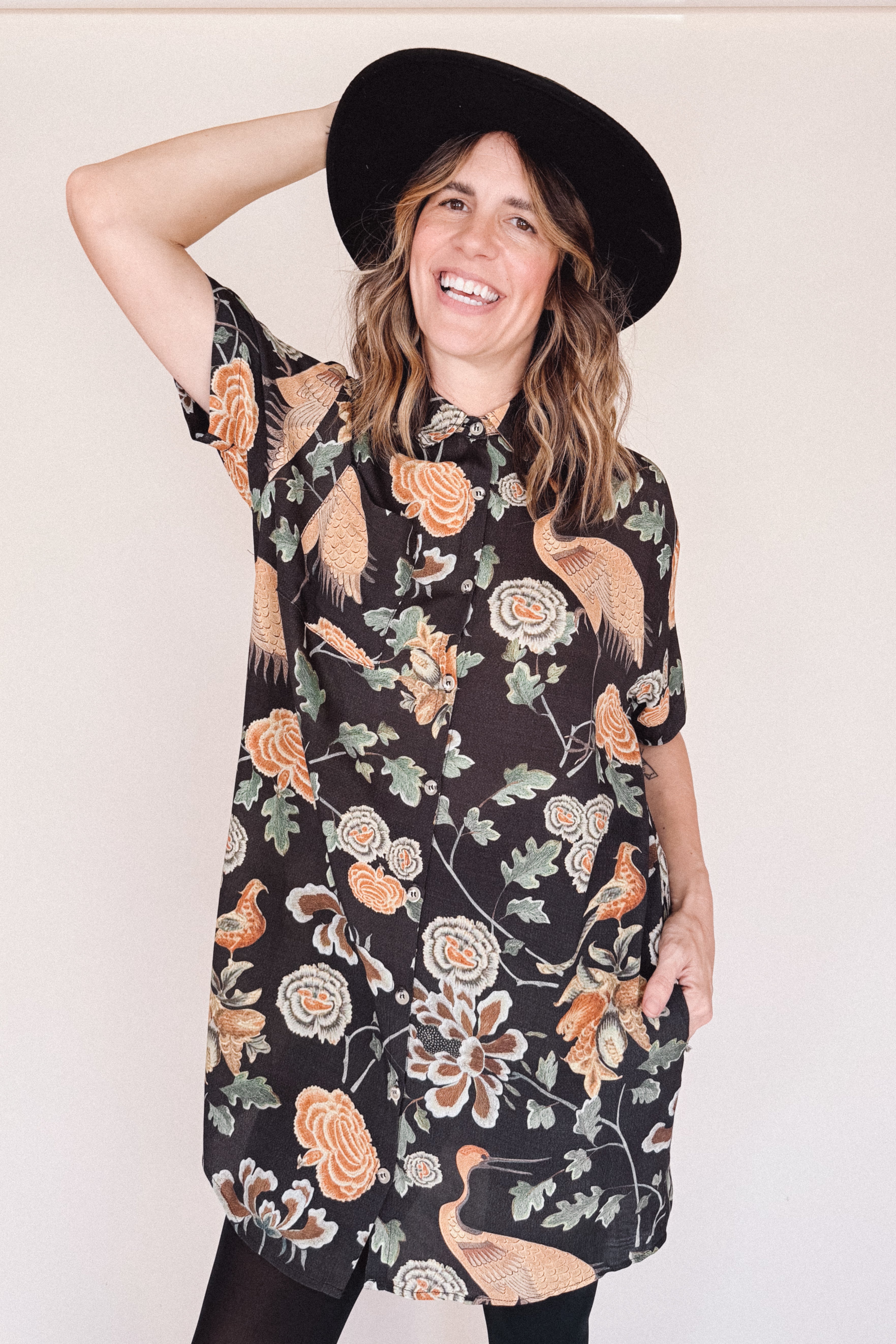 Woman wearing crane-print short sleeve button-up mini shirt dress with collar and side pockets, holding hat