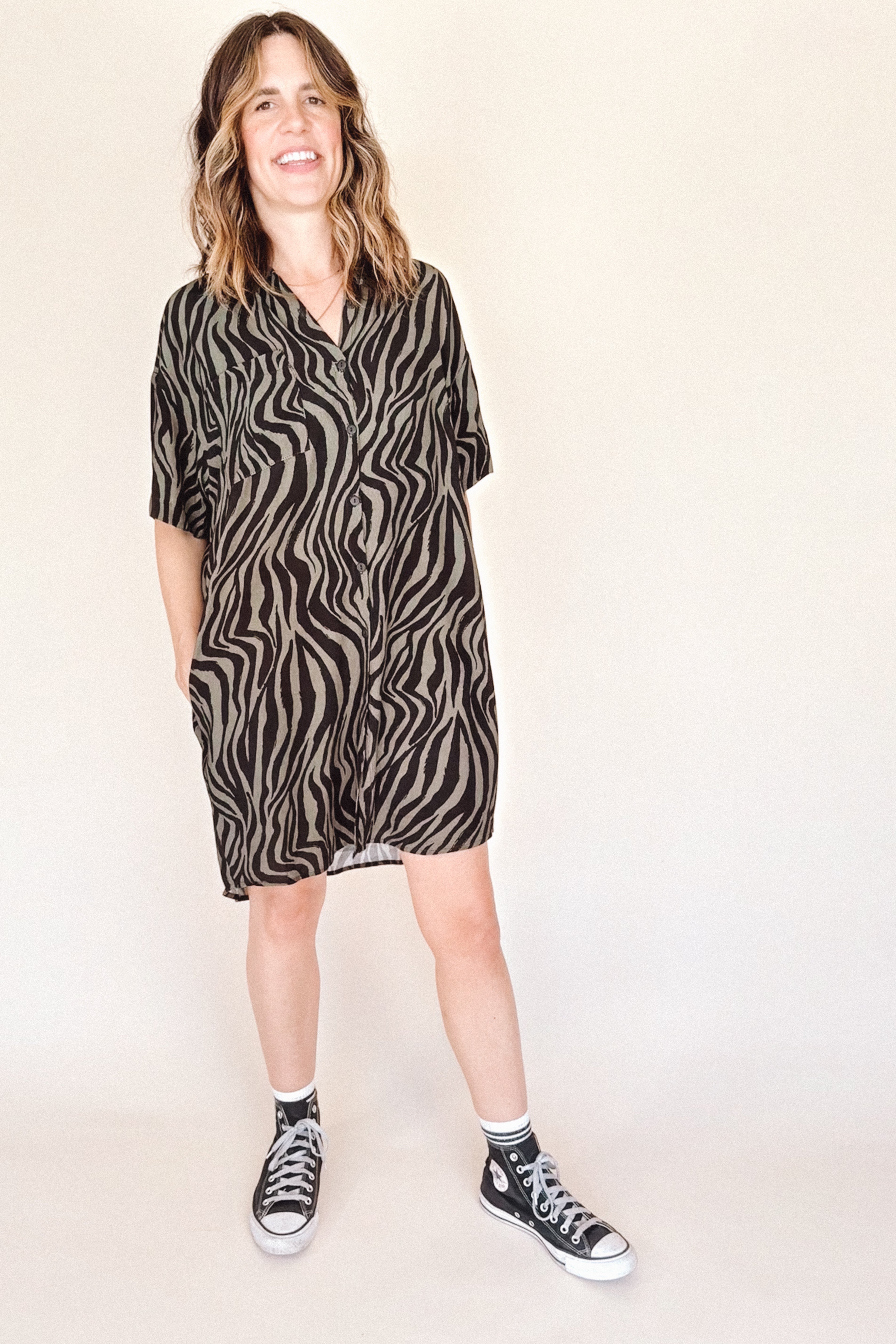woman wearing a black and olive zebra print button up mini shirt dress with black high-top sneakers on a beige background