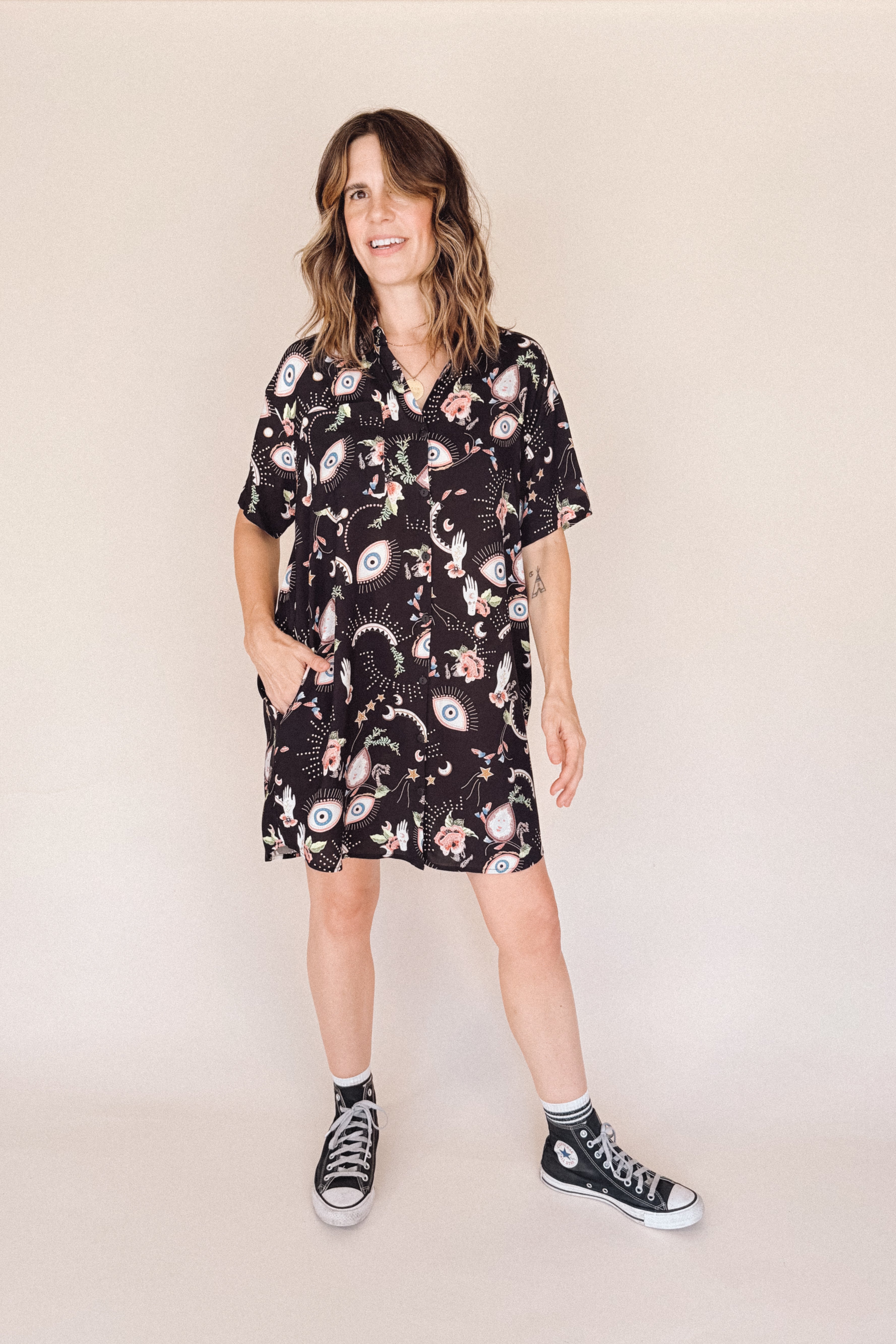Woman wearing a black eye printed shirt dress with black high-top sneakers on a beige background