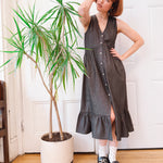 Woman in a gray dress standing next to a potted plant indoors.