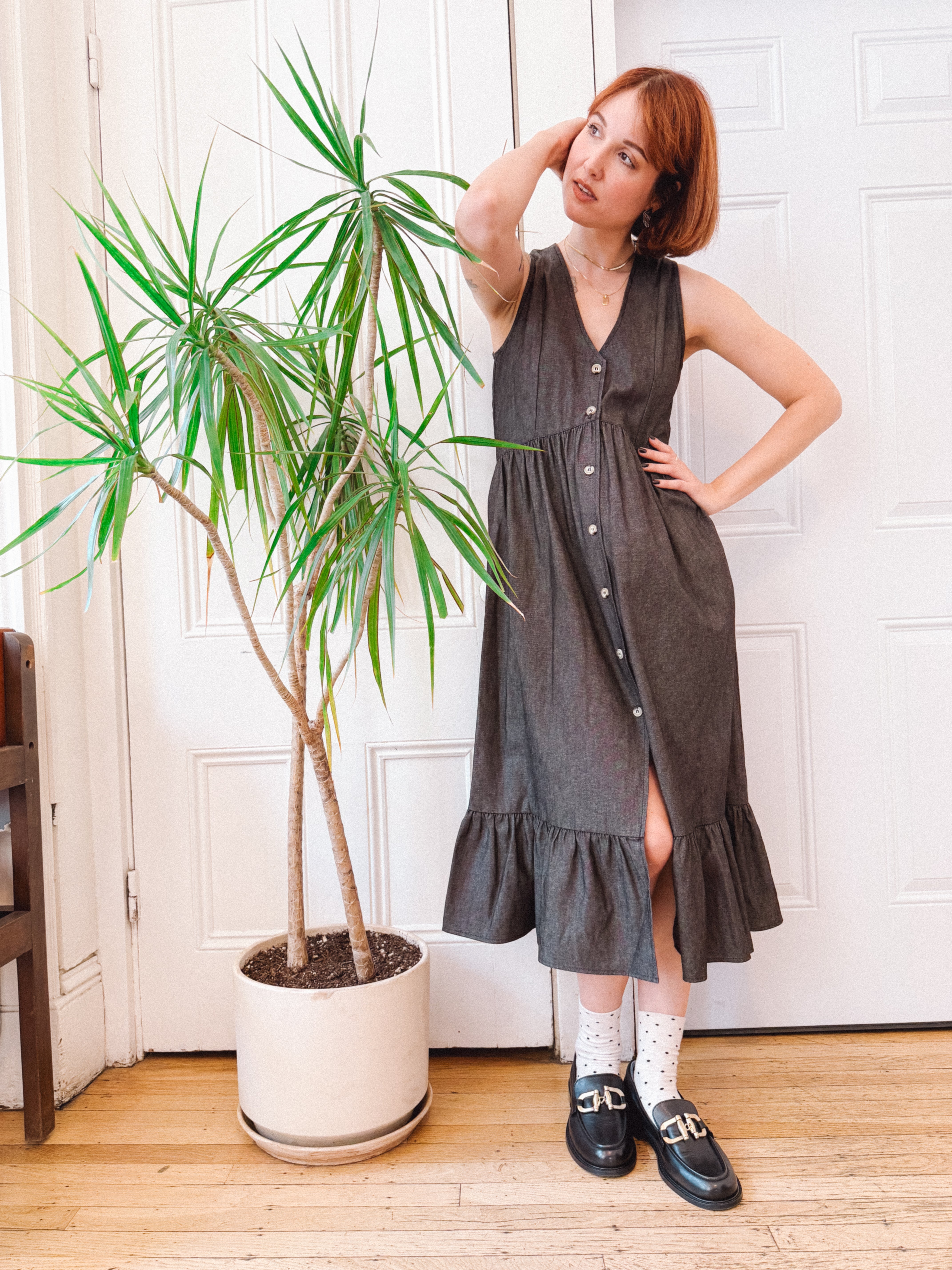 Woman in a gray dress standing next to a potted plant indoors.