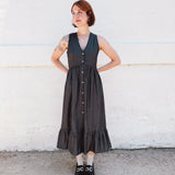 Woman wearing a black sleeveless dress with buttons against a white wall.