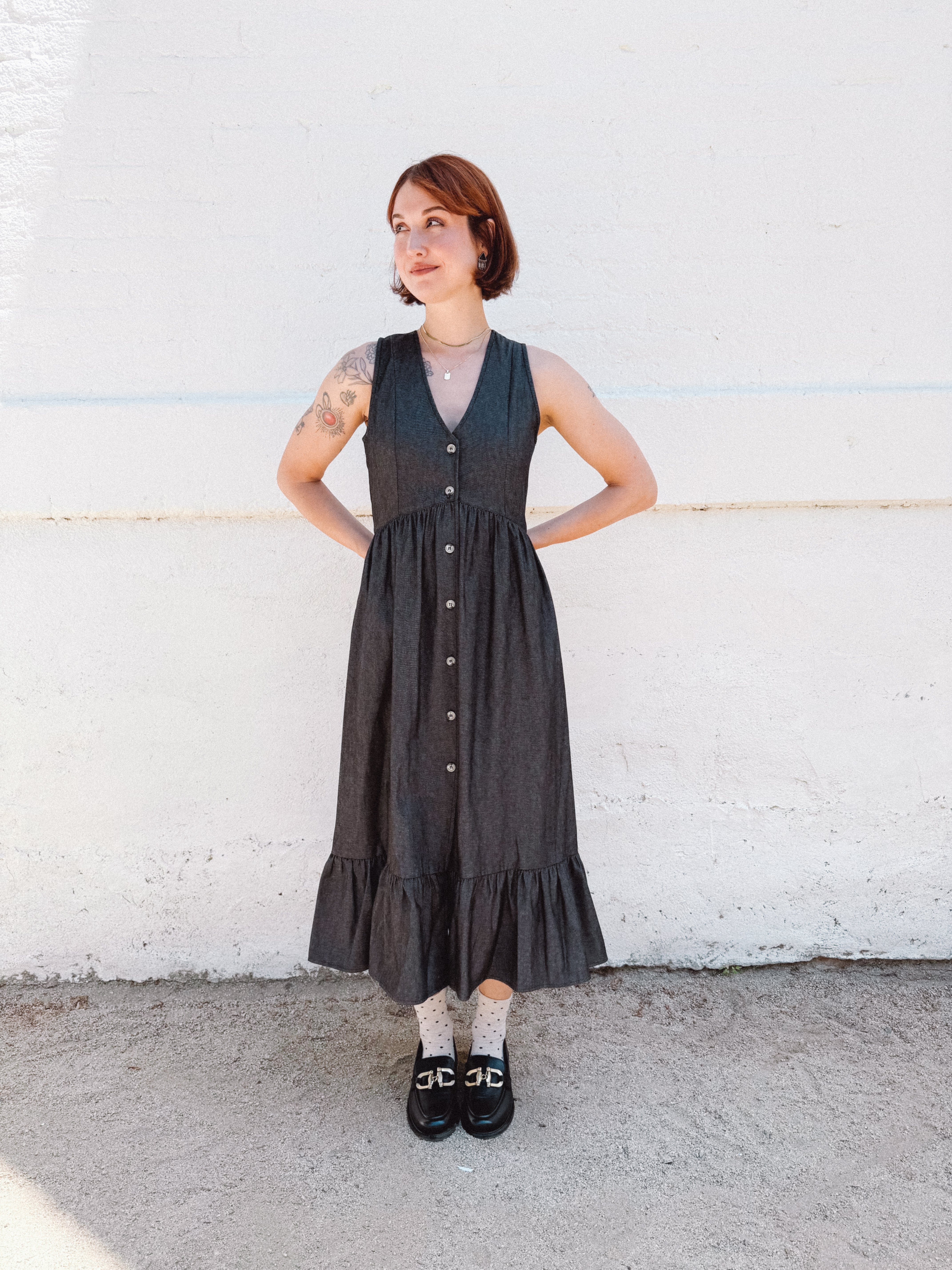 Woman wearing a black sleeveless dress with buttons against a white wall.