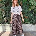 Woman wearing a eyeball printed crop white t-shirt, a brown and black zebra-striped skirt, and a beige cap, standing in front of green foliage.